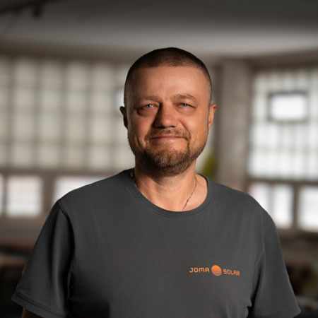 Man with short hair and beard wearing a dark gray Joma Sochr shirt smiling against a softly lit background.