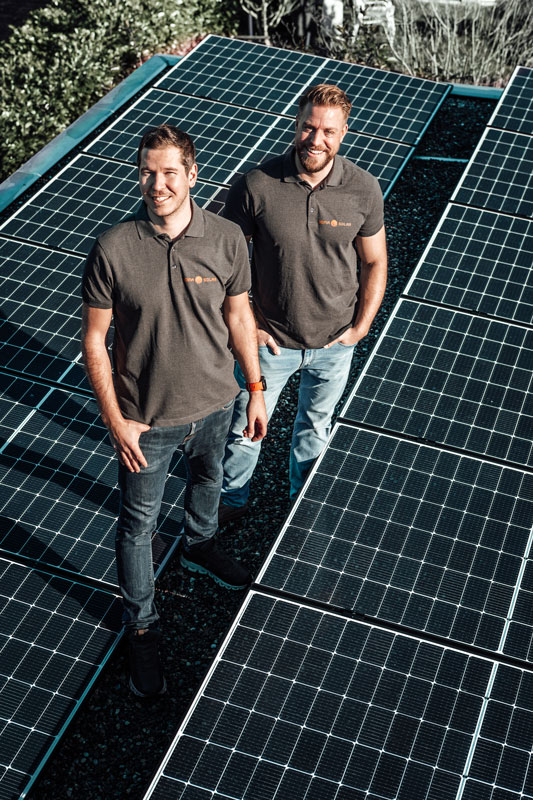 Two men in matching gray polo shirts standing and smiling between rows of solar panels outdoors.