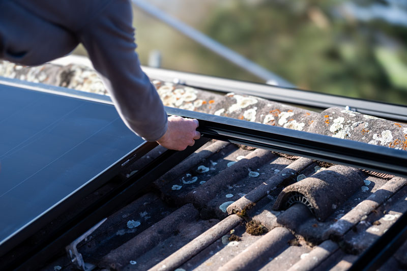 Person's hand installing or adjusting a solar panel frame on a tiled roof with lichen and moss.