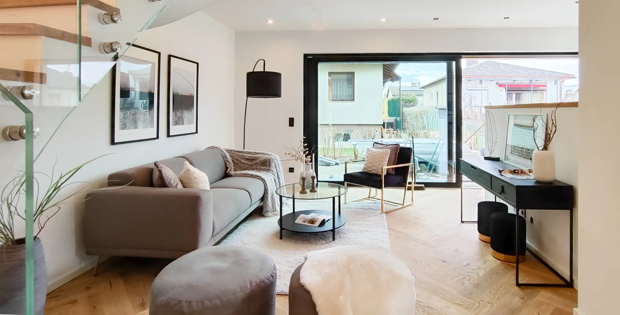 Modernes Wohnzimmer mit grauem Sofa, schwarzen Sessel, Glas-Couchtisch und großem Fenster mit Blick auf den Garten.