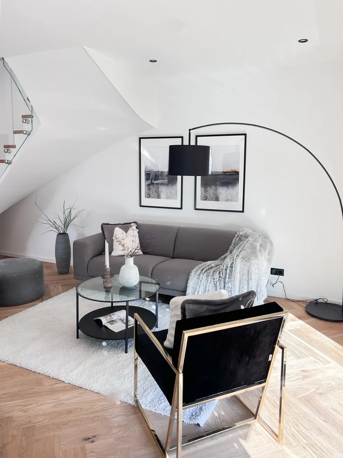 Modernes Wohnzimmer mit grauem Sofa, schwarzem Sessel mit goldfarbenem Rahmen, rundem Glastisch und großem Bogenstehlampe.