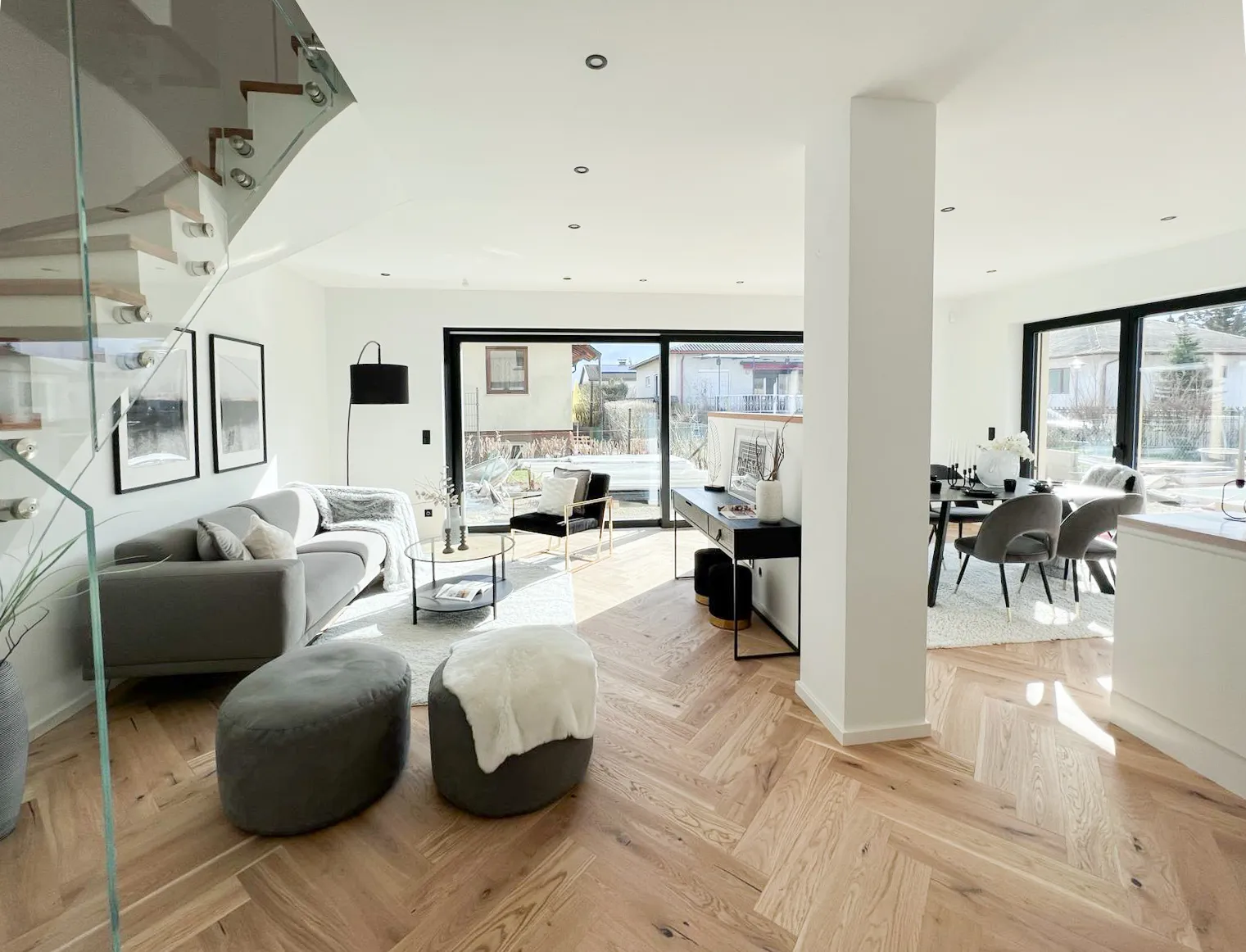 Heller, moderner Wohn- und Essbereich mit Parkettboden, grauem Sofa, Sesseln, Glas-Treppen-Geländer und großen Fenstern.
