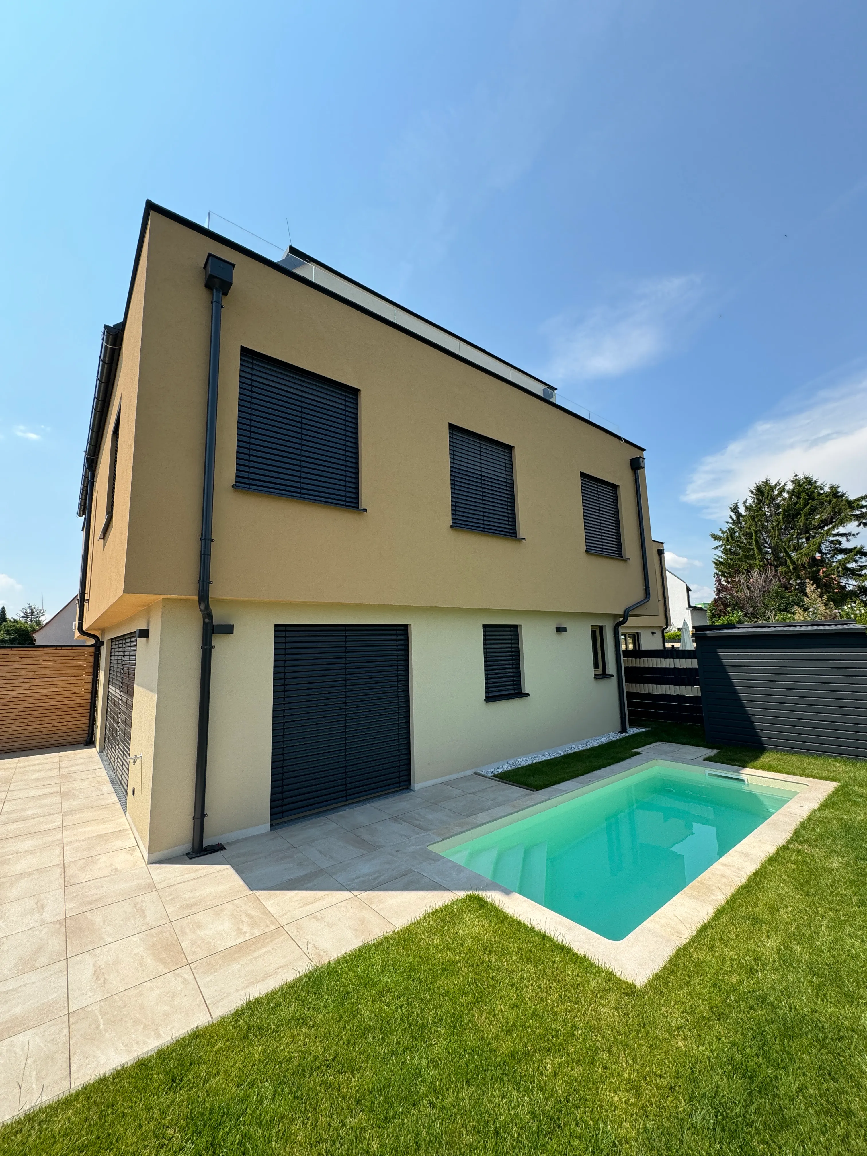 Modern zweistöckiges Haus mit beige Fassade, schwarzen Fensterläden, kleinem rechteckigen Pool und grünem Rasen im Garten.