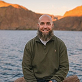 Smiling bald man with a beard sitting near a lake with brown mountains in the background.