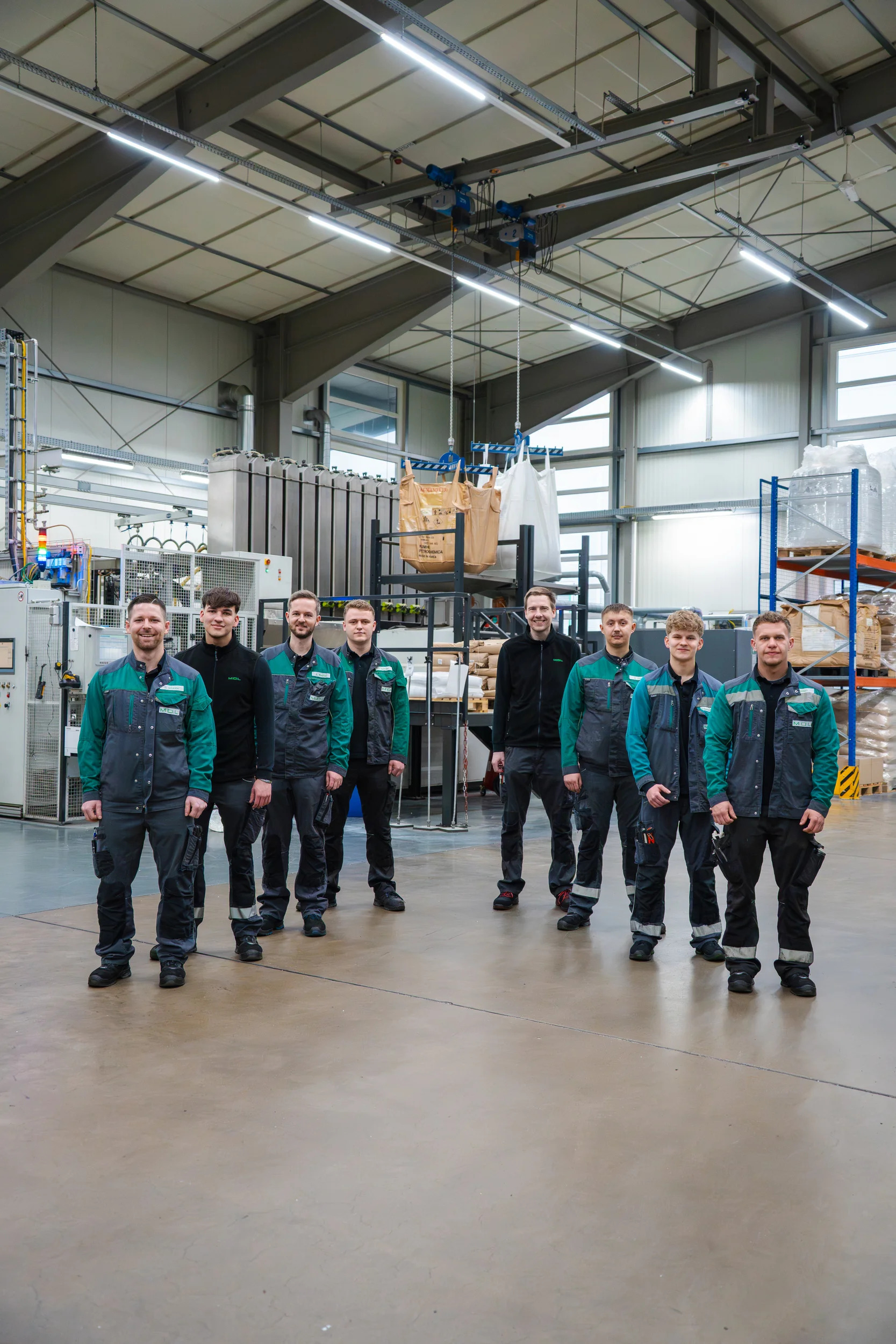 Acht Männer in Arbeitskleidung, die in einer modernen Fabrikhalle stehen und lächeln.