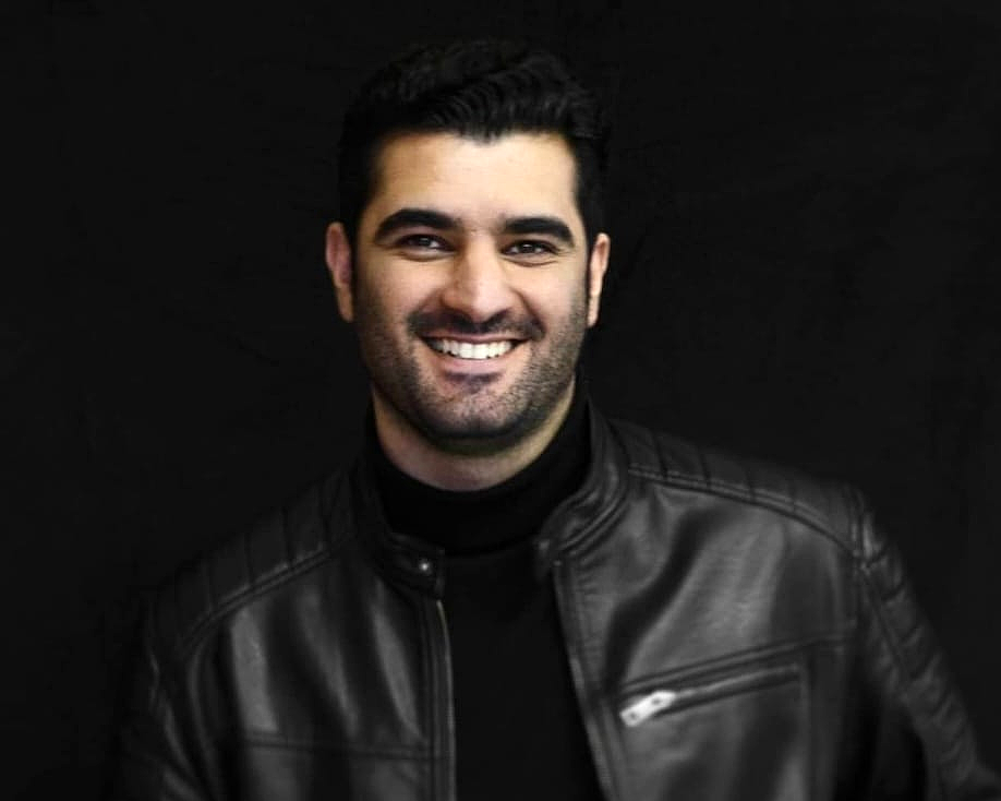 Smiling man with dark hair wearing a black leather jacket and black shirt against a black background.