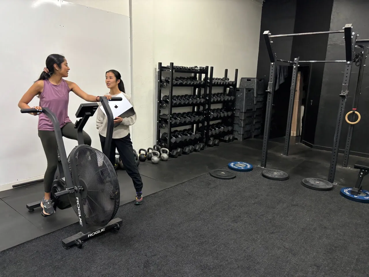 Two women in a gym, one using a Rogue air bike and the other holding a laptop while talking.