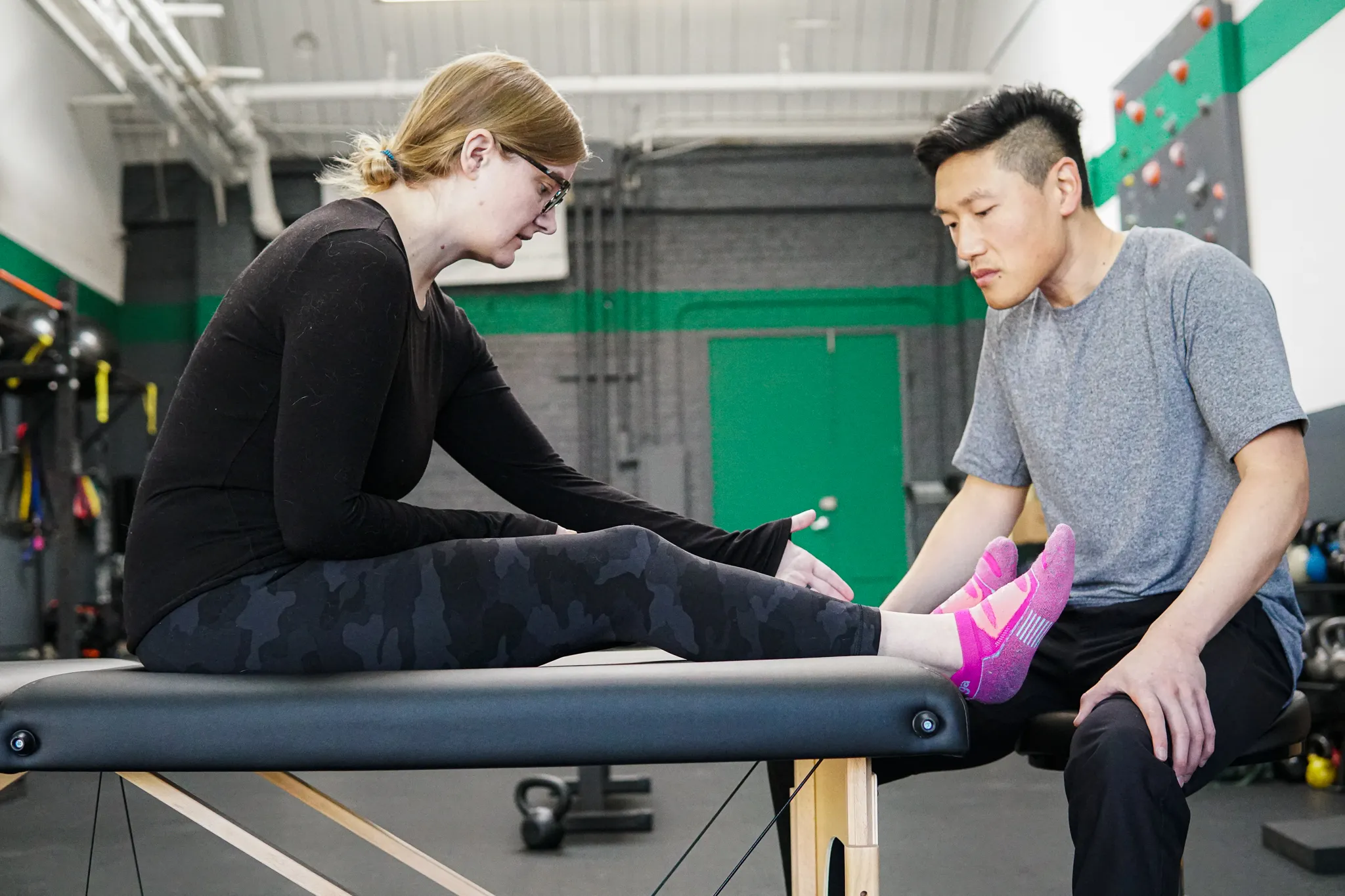 Physical therapist assisting a woman in stretching her legs on a treatment table in a gym.