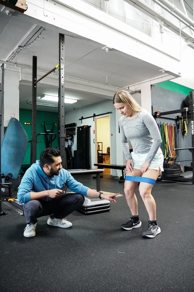 Fitness trainer crouching and pointing while coaching woman with resistance band around thighs in gym.
