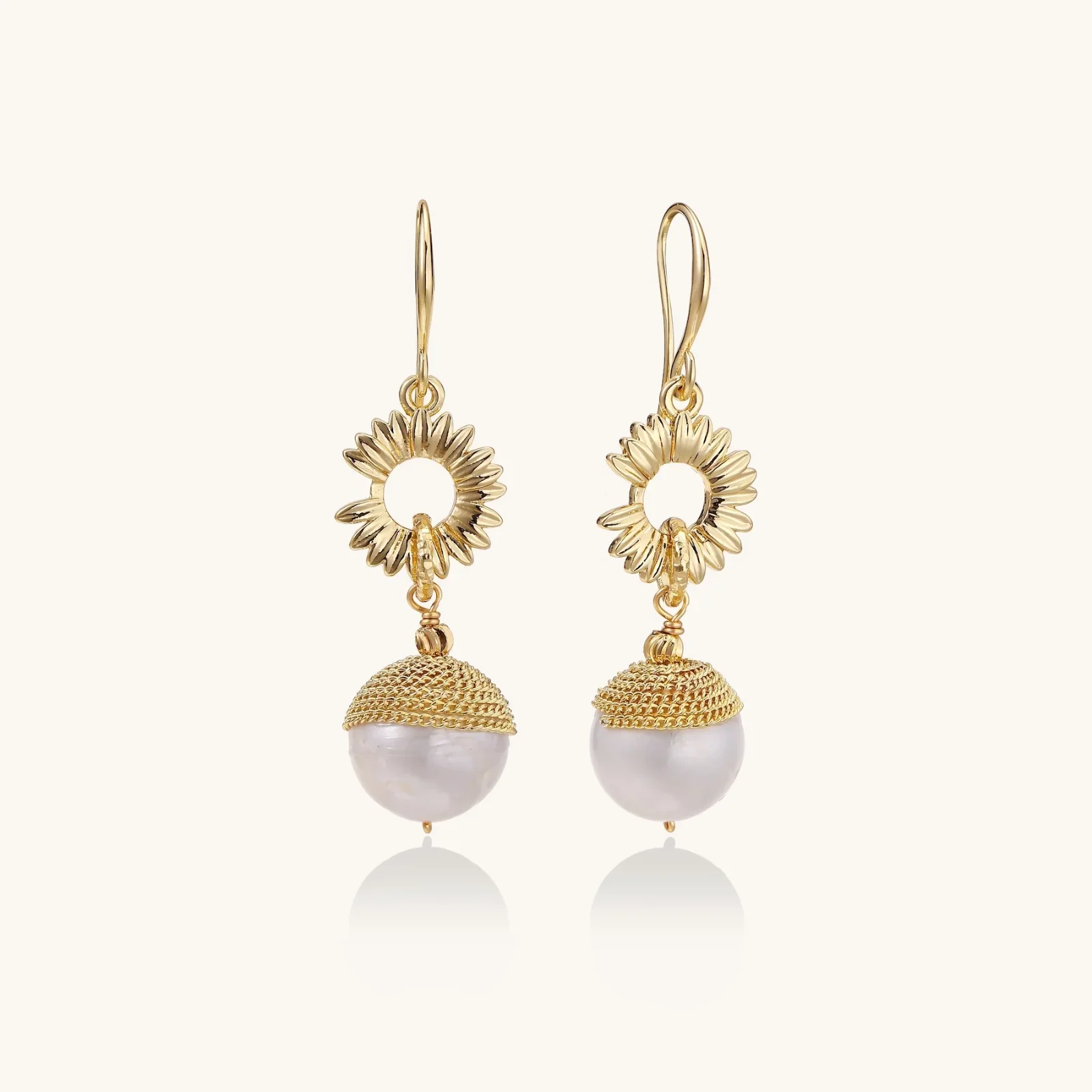Gold sunburst charm earrings with baroque pearl drops on French hooks against cream background