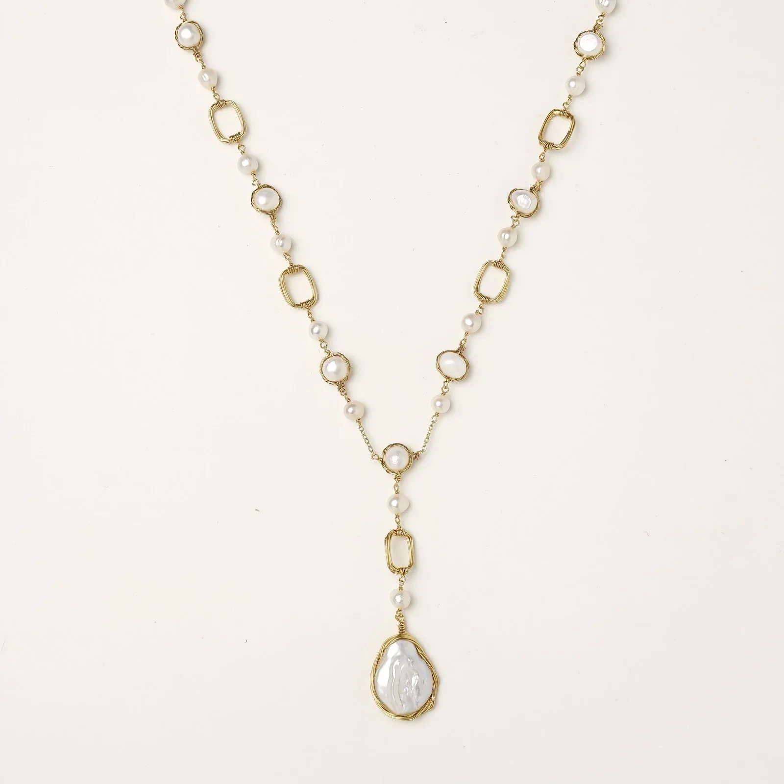 Y-shaped gold necklace with alternating geometric links, freshwater pearls, and baroque pearl pendant on cream background