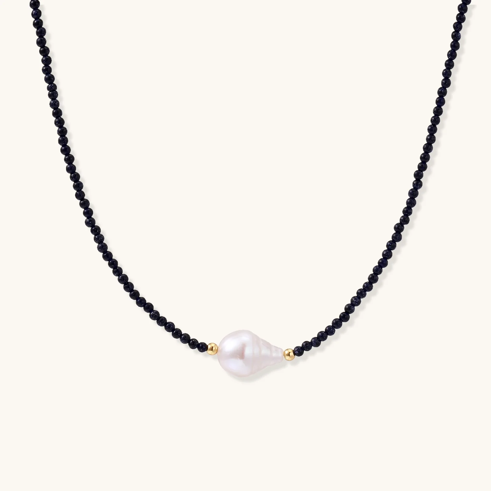 Delicate necklace with black blue sandstone beads and baroque pearl center on white background with gold accents.