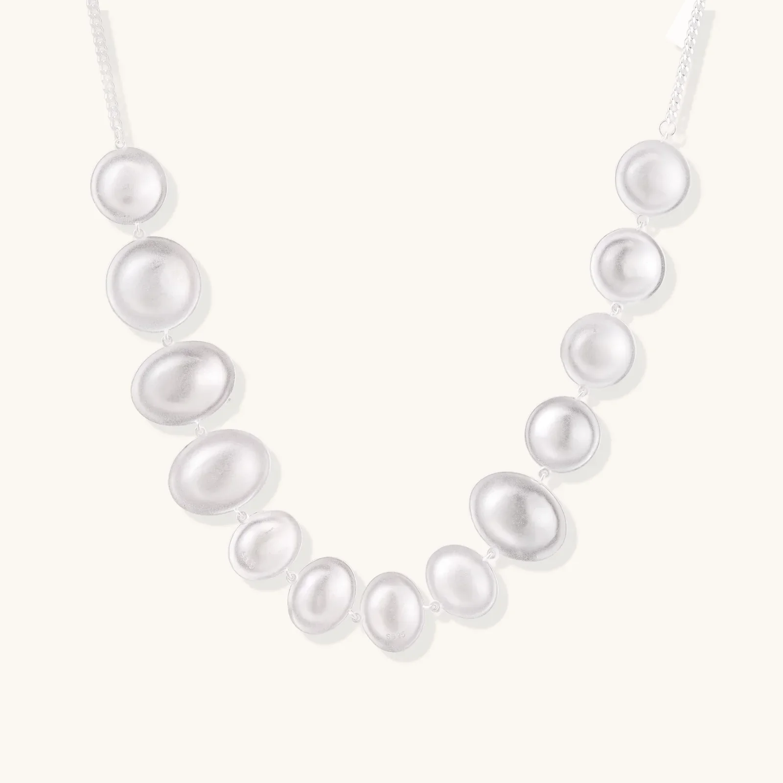 Graduated pearl-finish silver discs forming an organic bubble bib necklace on delicate chain against cream background.