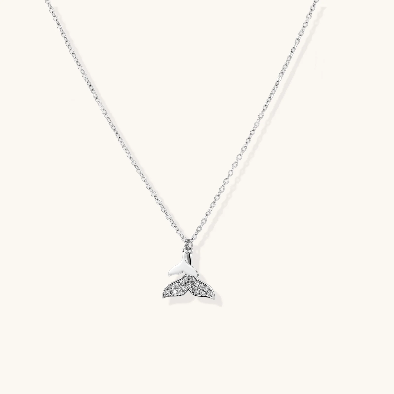 Delicate white gold plated whale tail pendant with pavé crystals on silver chain against cream background.