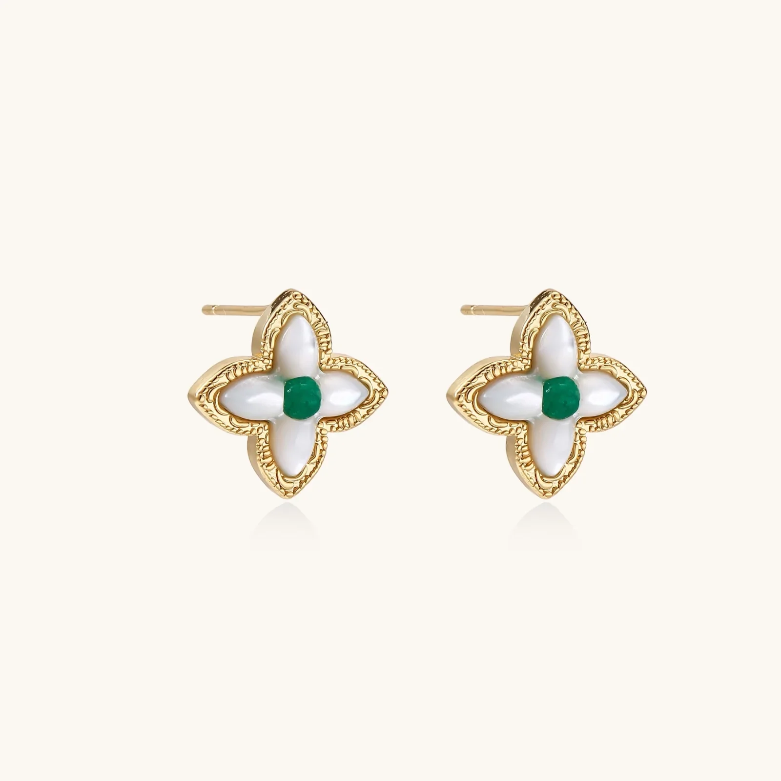 Gold quatrefoil stud earrings with white mother-of-pearl petals and emerald green gemstone center on beige background.