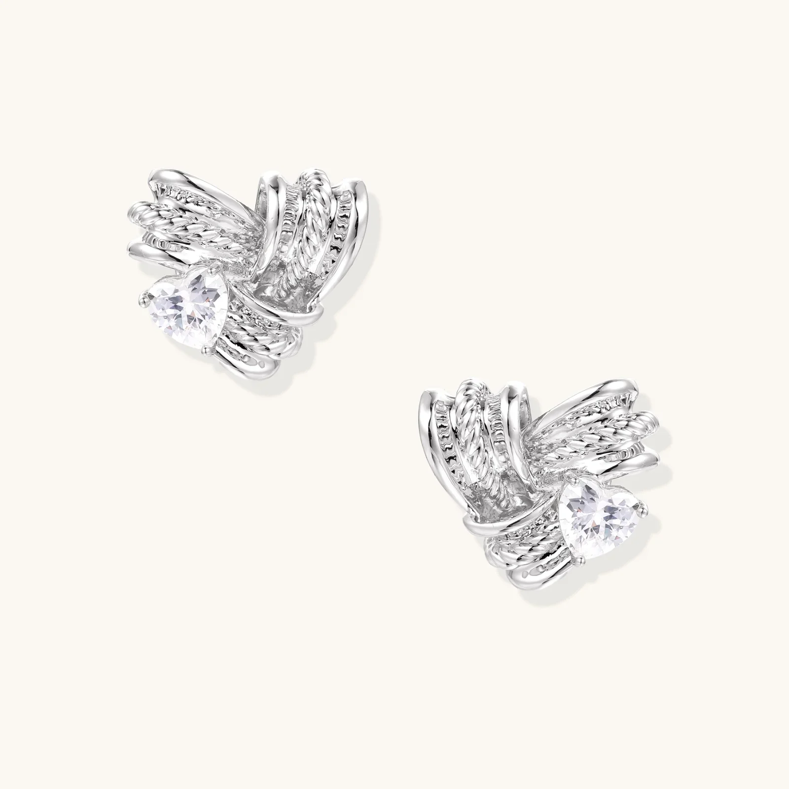 Pair of sculptural white gold-plated stud earrings with twisted ribbon design and sparkling zirconia stones on cream background