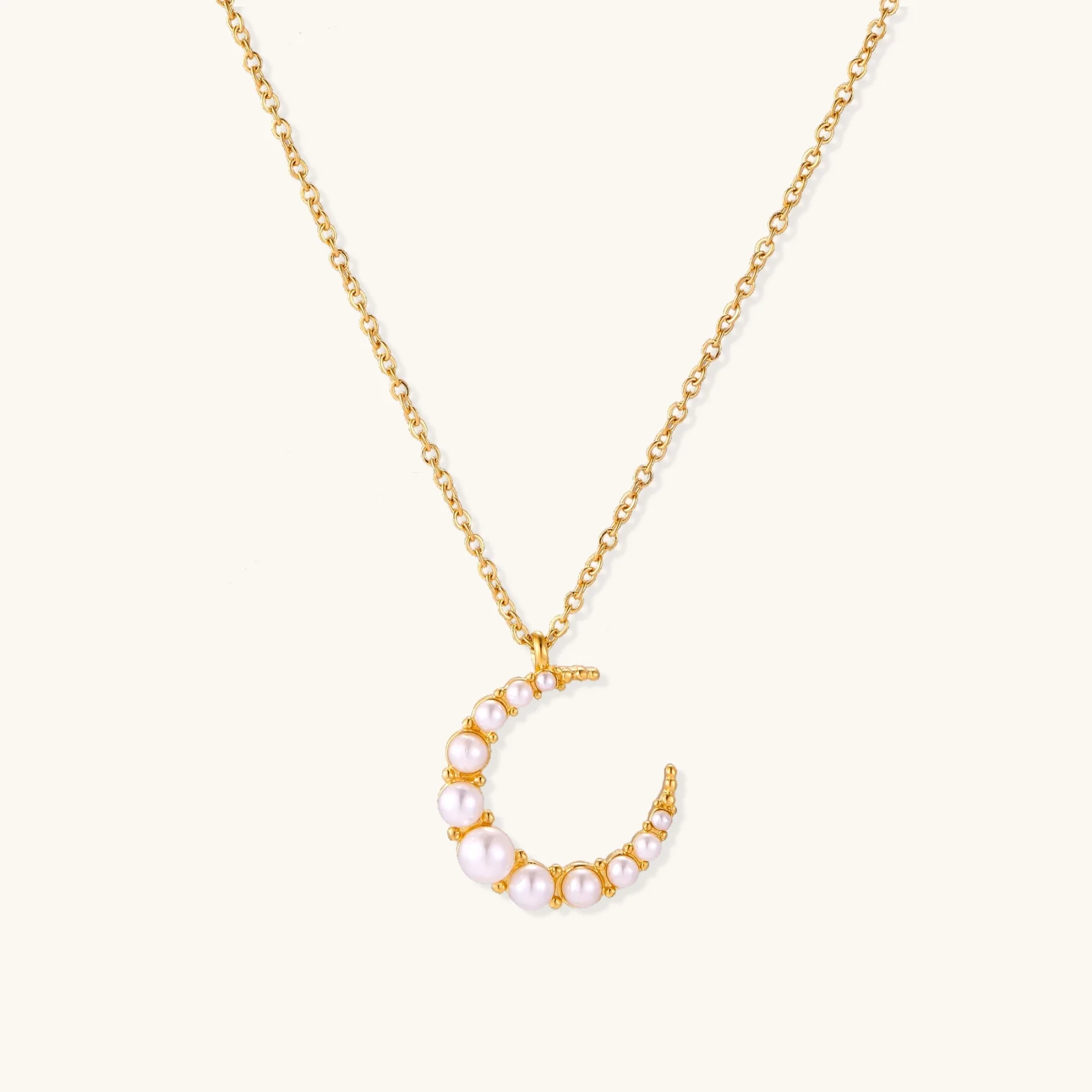 Gold crescent moon pendant with graduated white pearls on delicate chain against cream background