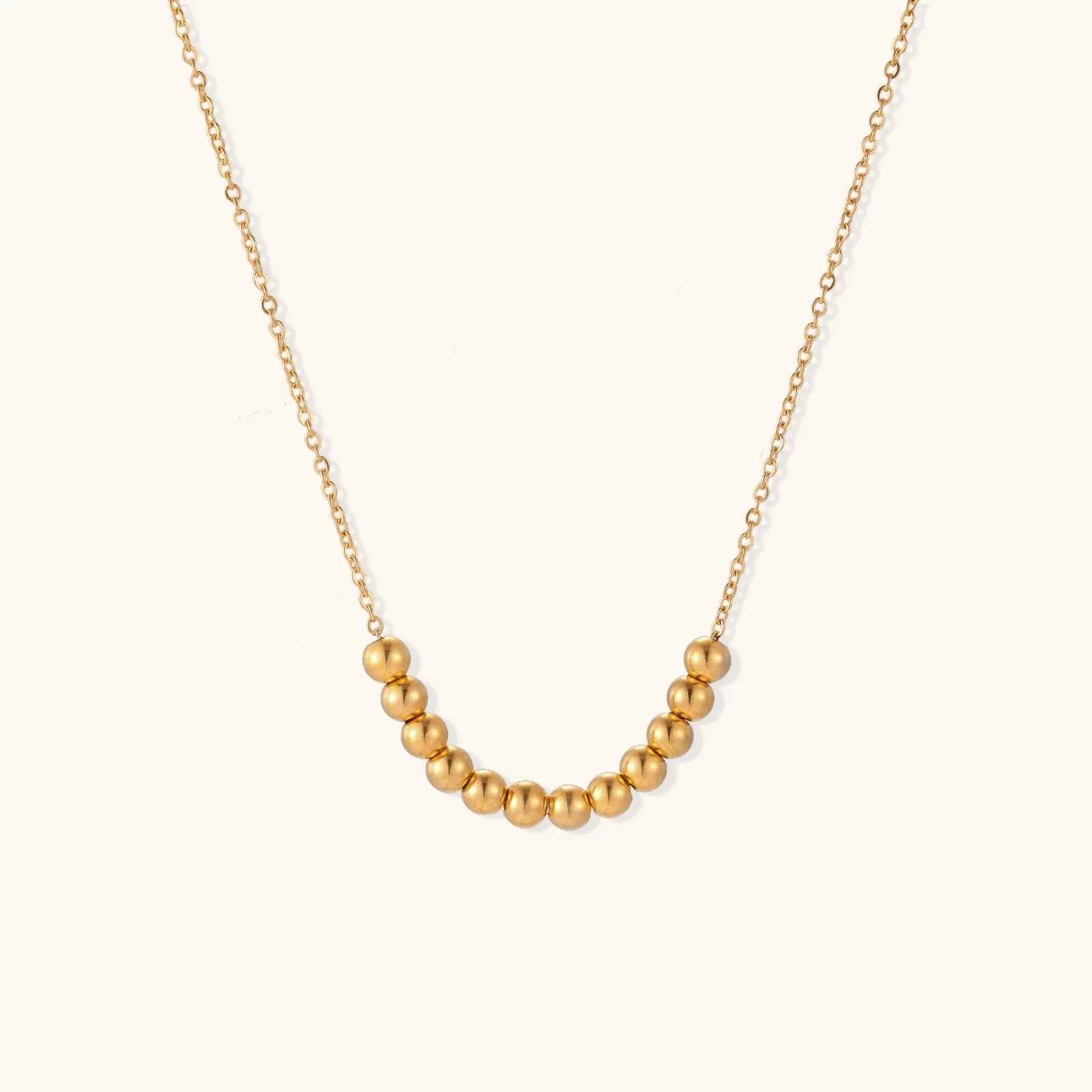 Delicate gold-plated necklace with graduated spherical beads forming gentle smile curve on beige background