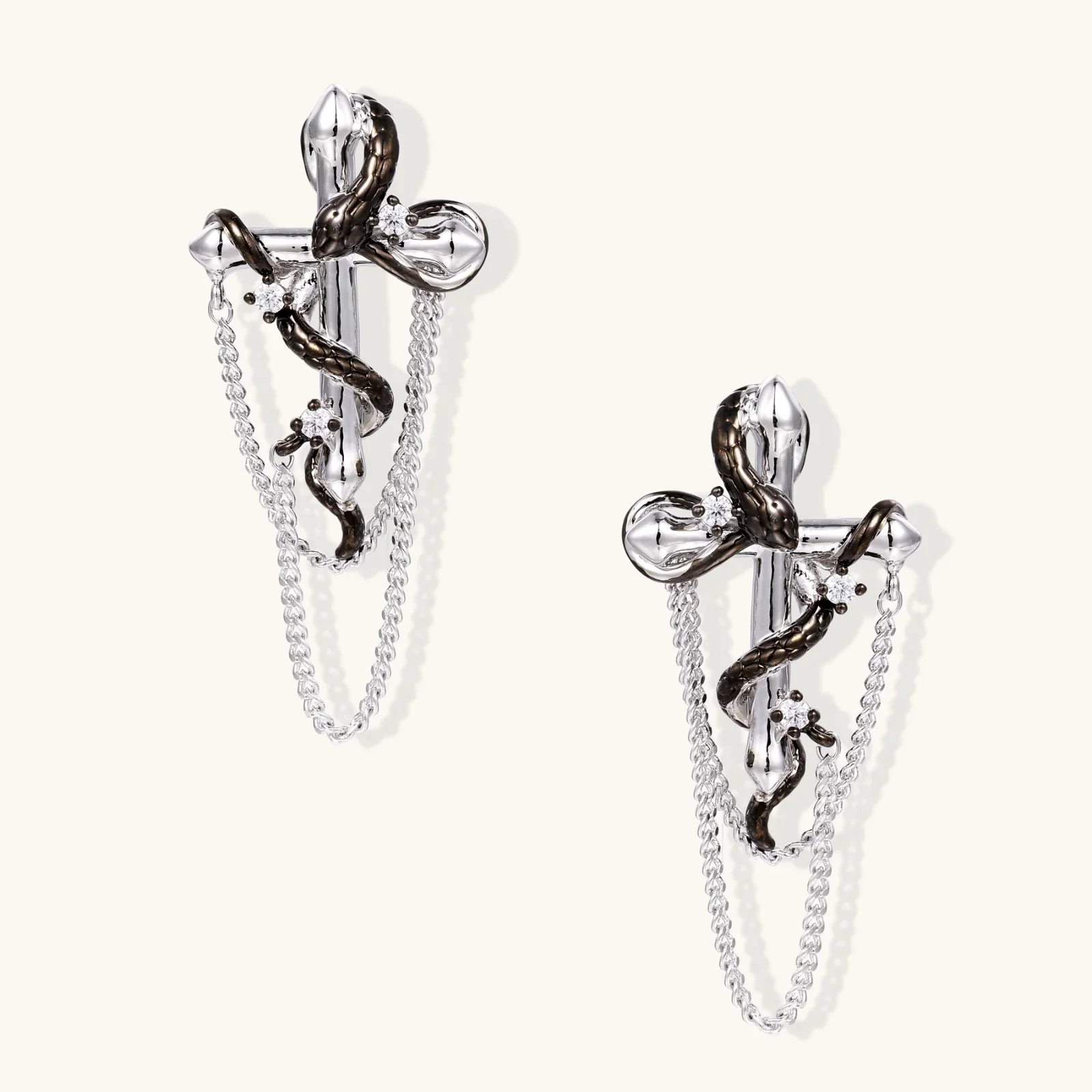 Silver dangle earrings with sculptural serpent coiled around cross shape and delicate chain accents on cream background