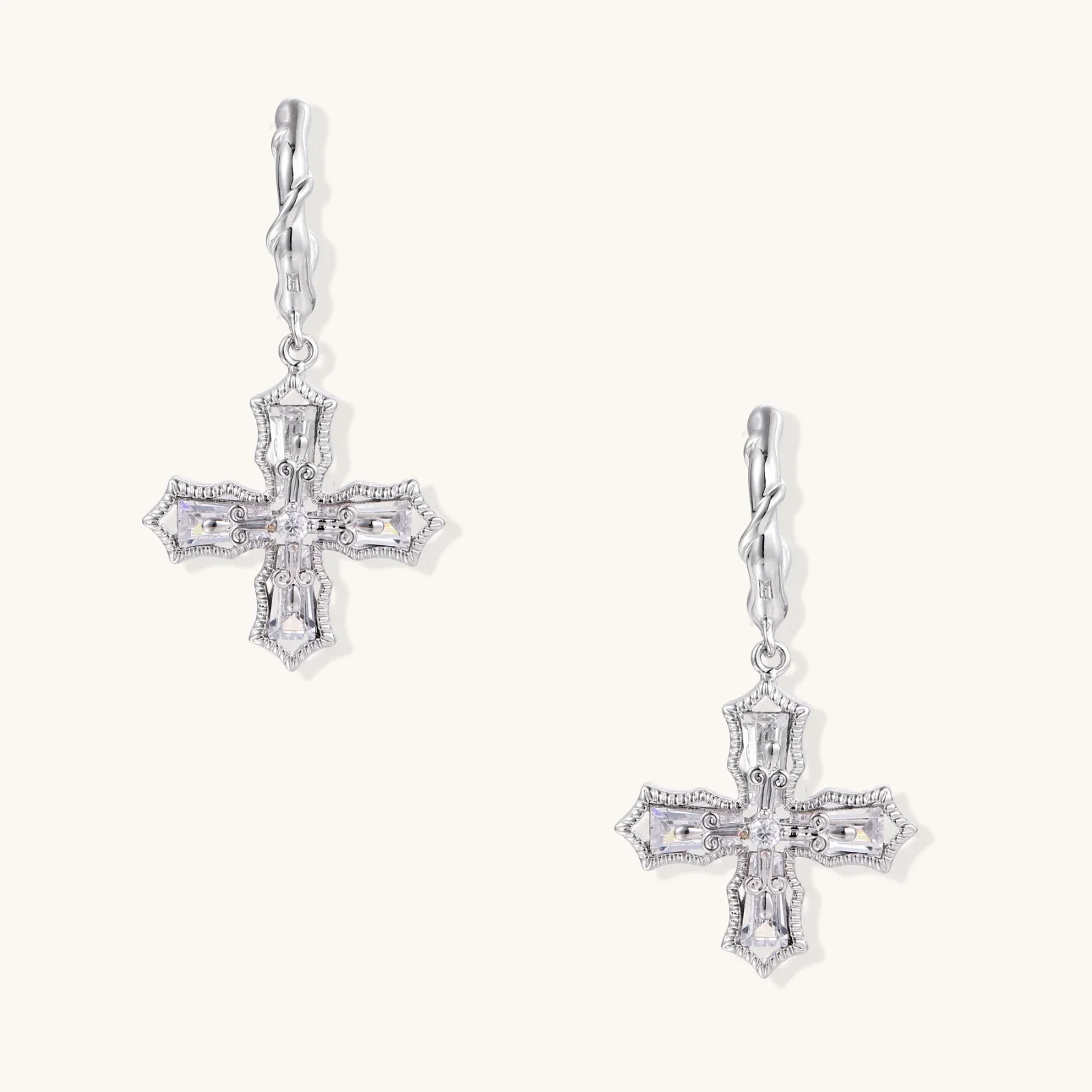 Ornate white gold-plated cross earrings with baguette-cut zirconia stones and twisted leverback hooks on beige background