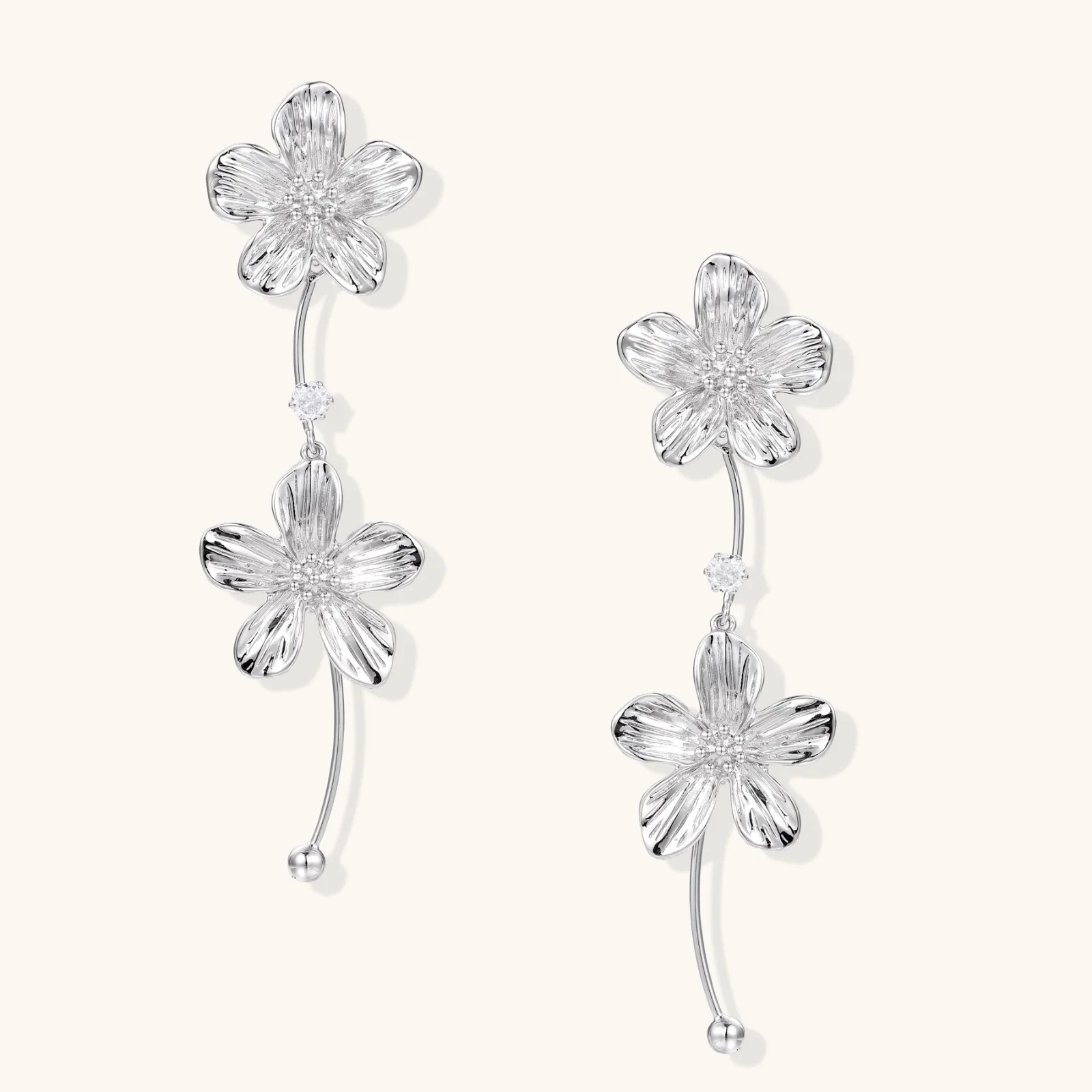 Silver-white asymmetric earrings with textured flower petals and crystal centers on cream background