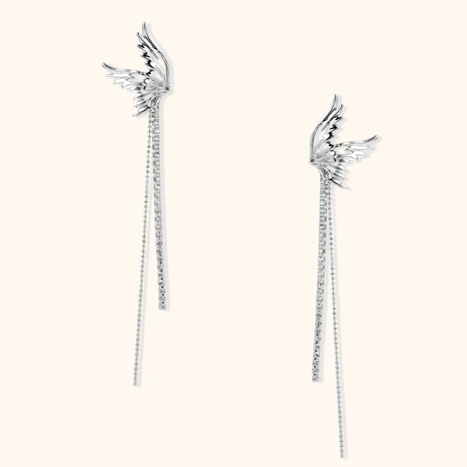 Pair of silver wing-shaped earrings with feather details and long sparkling crystal chain drops on cream background