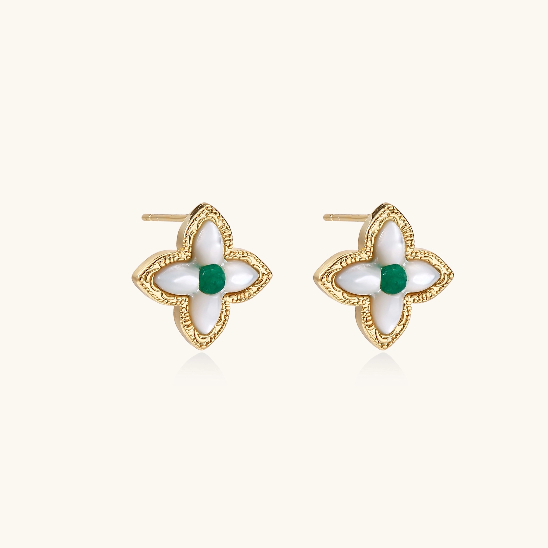 Gold quatrefoil stud earrings with white mother-of-pearl petals and emerald green gemstone center on beige background.