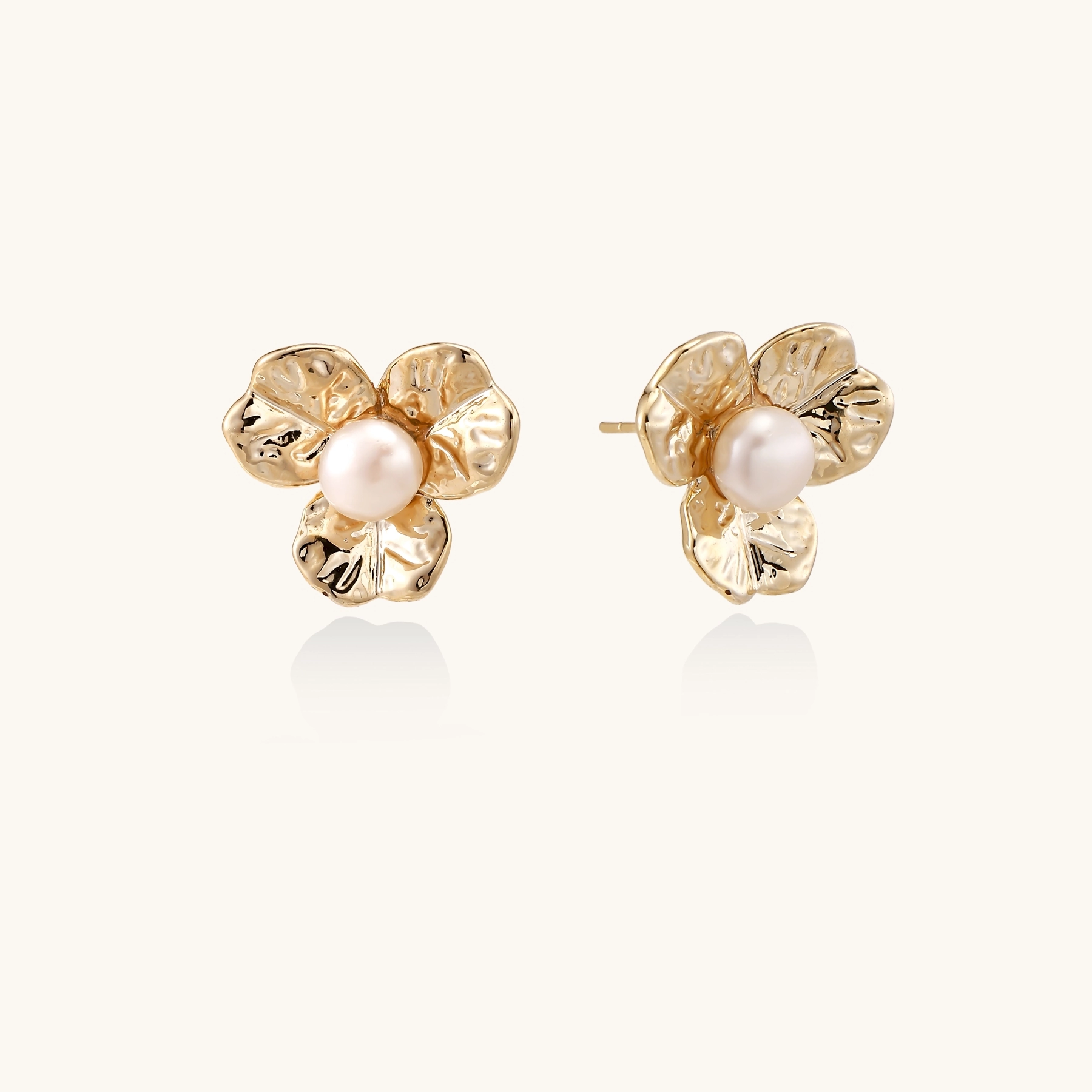 Gold-plated flower stud earrings with textured petals and centered freshwater pearl on beige background