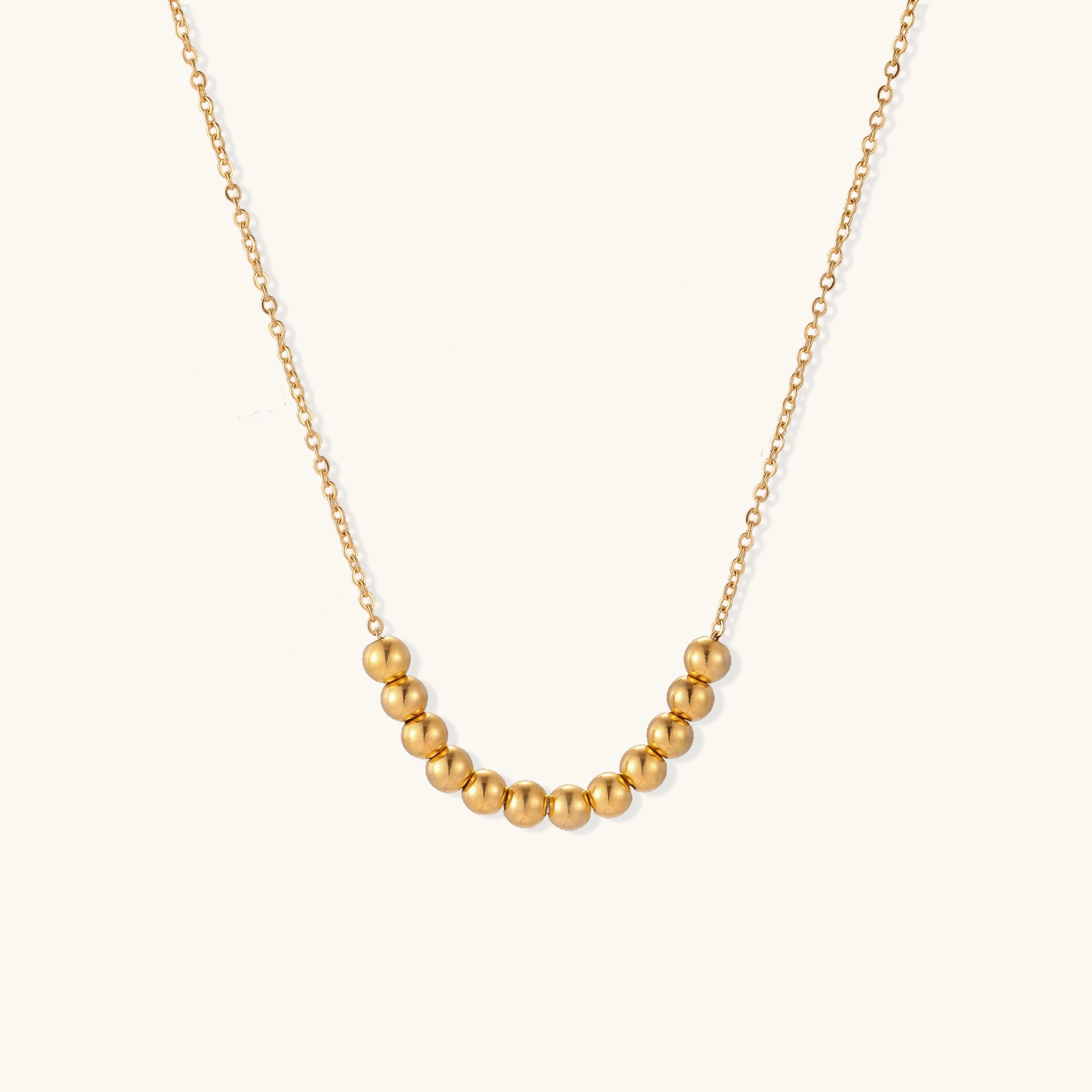 Delicate gold-plated necklace with graduated spherical beads forming gentle smile curve on beige background