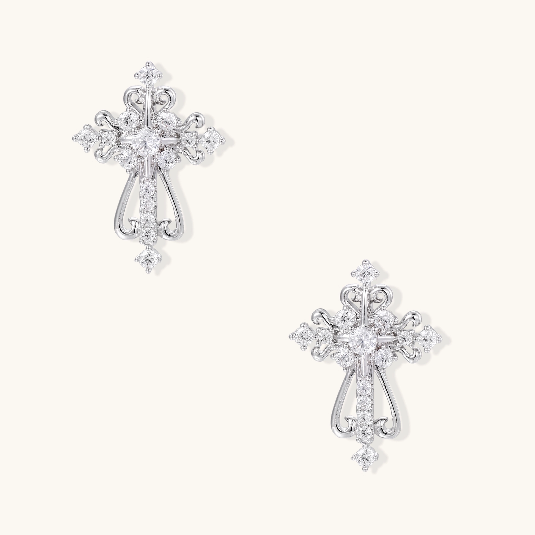 White gold-plated baroque cross studs with zirconia stones and ornate scrollwork on cream background.
