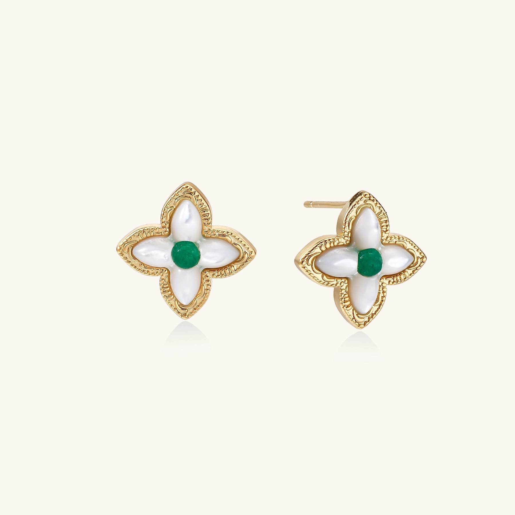 Gold quatrefoil stud earrings with white mother-of-pearl petals and emerald green gemstone center on beige background.