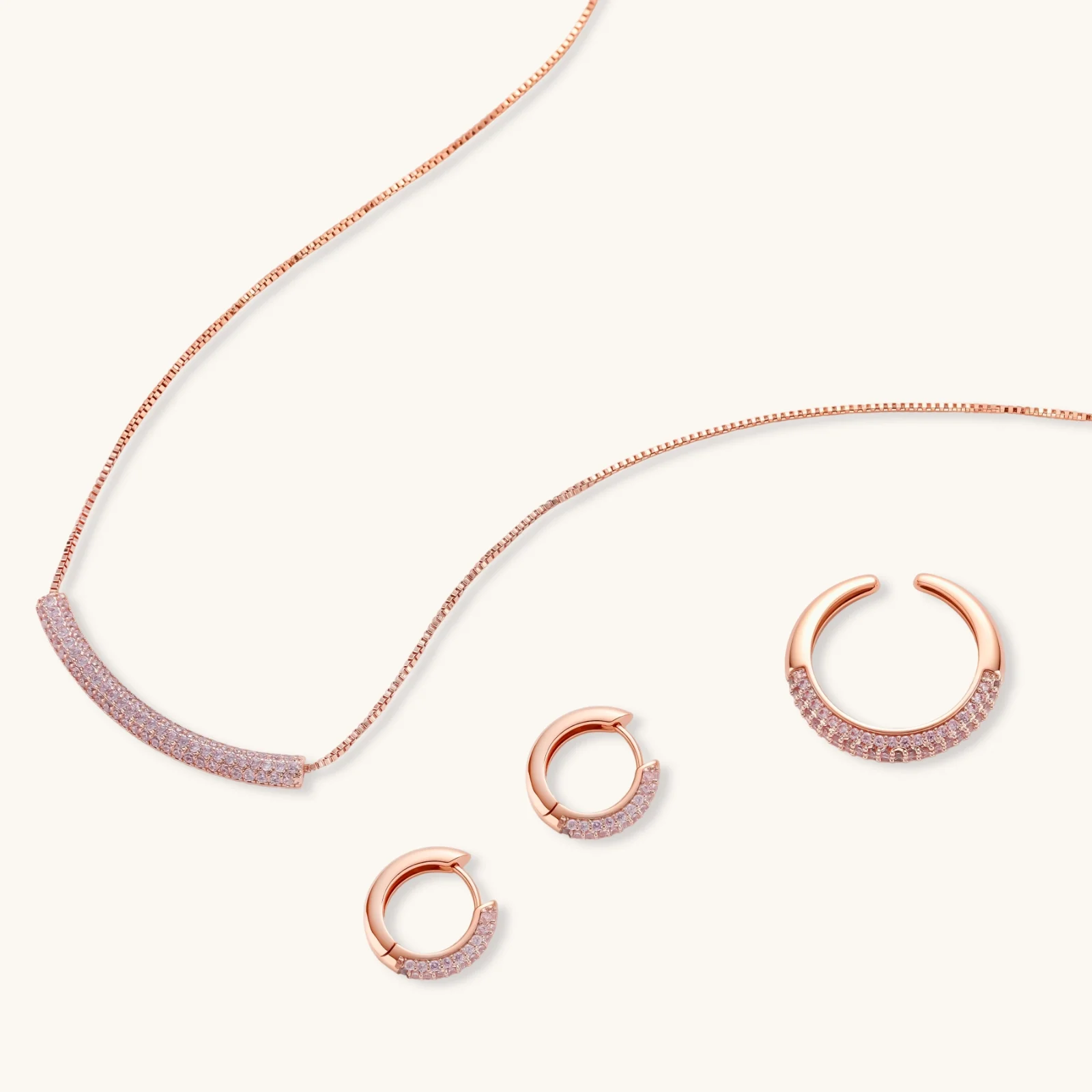 Rose gold jewelry set with pavé cubic zirconia stones including curved necklace, hoop earrings, and adjustable ring on cream background.