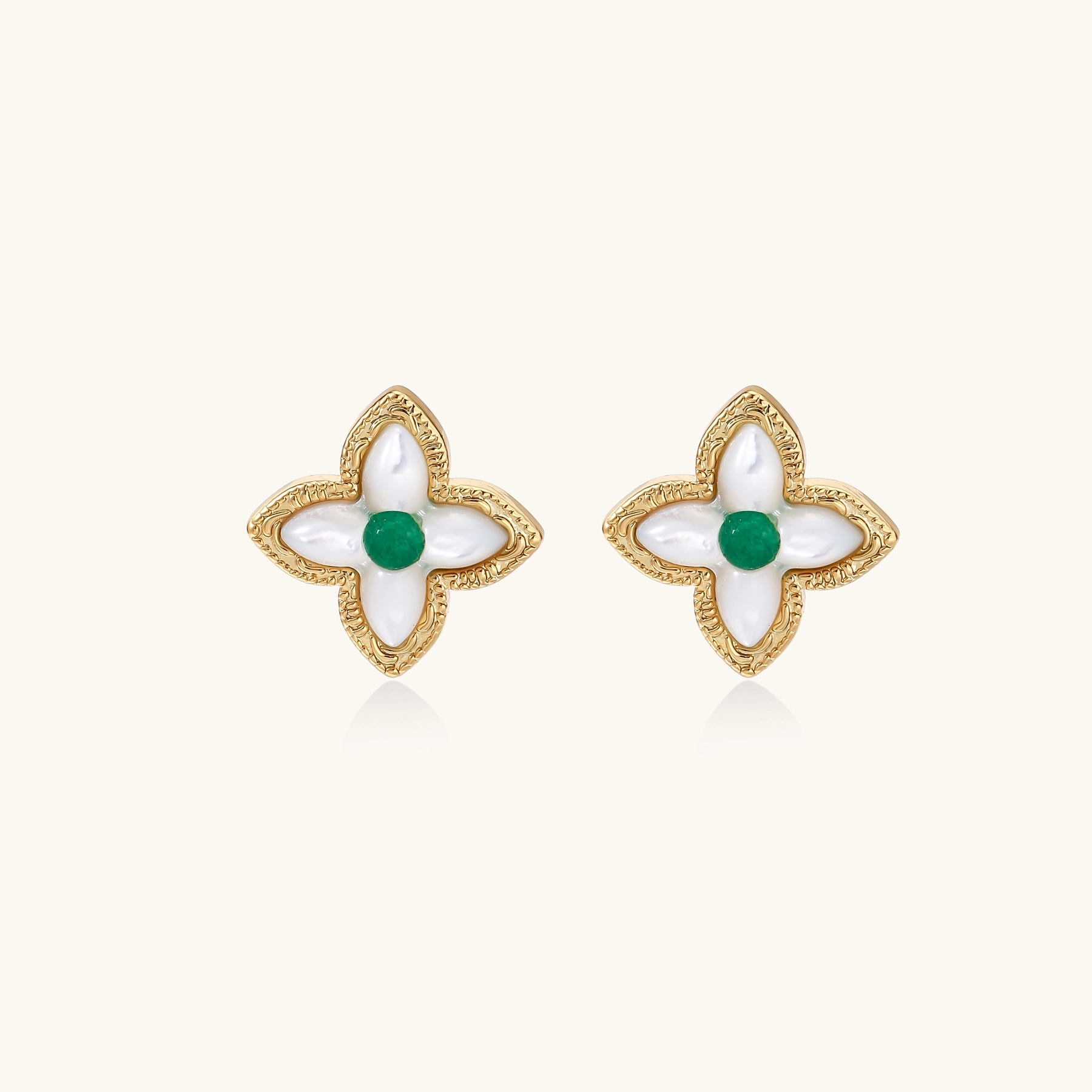 Gold quatrefoil stud earrings with white mother-of-pearl petals and emerald green gemstone center on beige background.