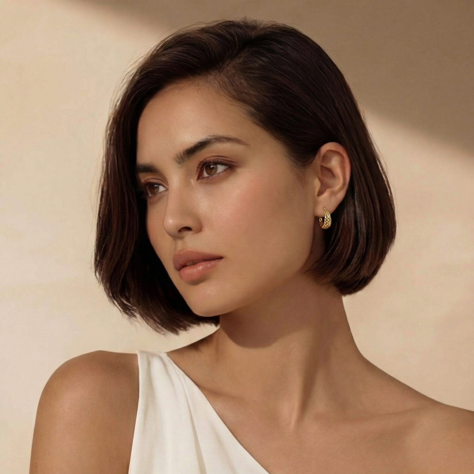 Close-up of small gold-plated hoop earrings with intricate honeycomb cutout pattern on cream background.