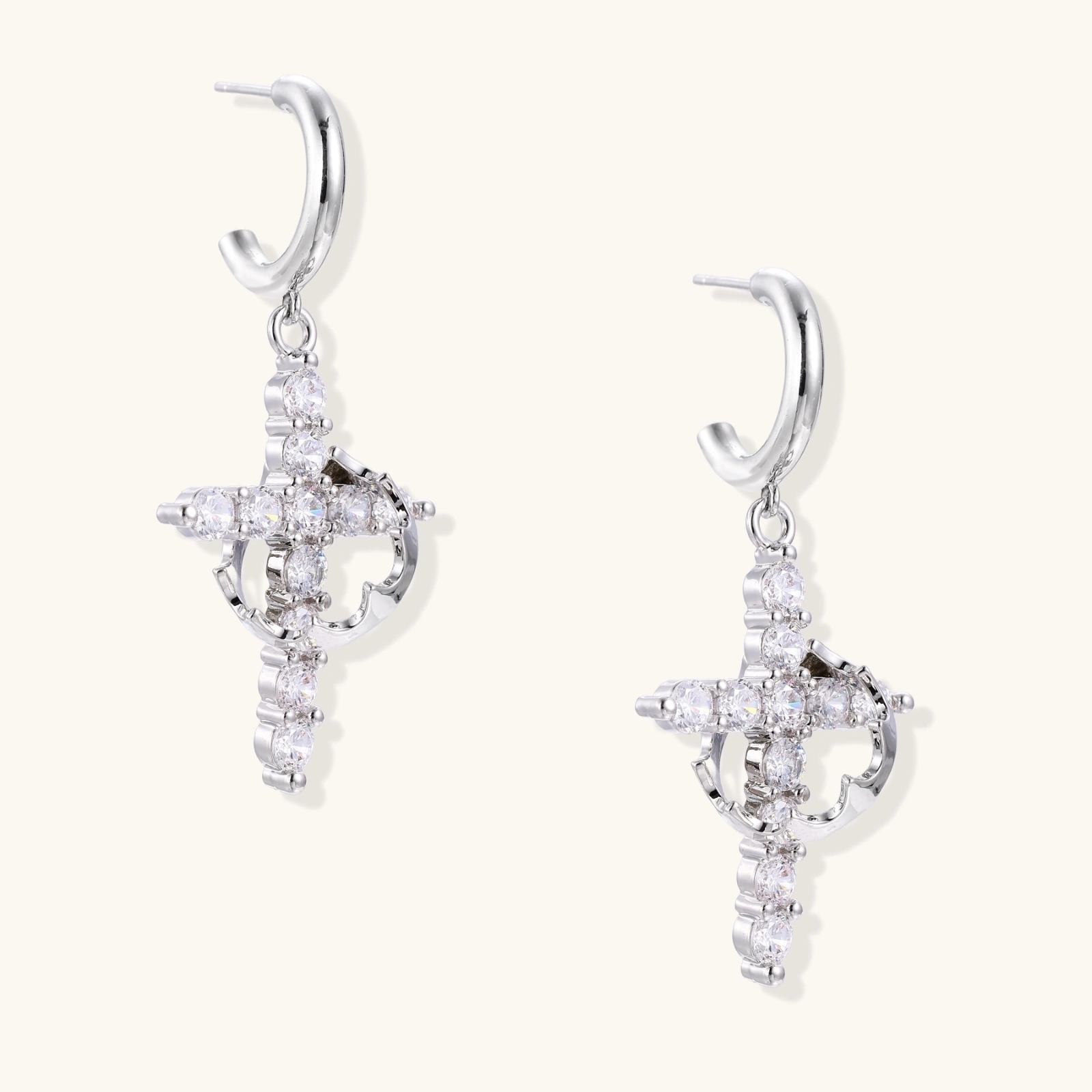 White gold-plated hoop earrings with sparkling zirconia cross charms dangling against cream background