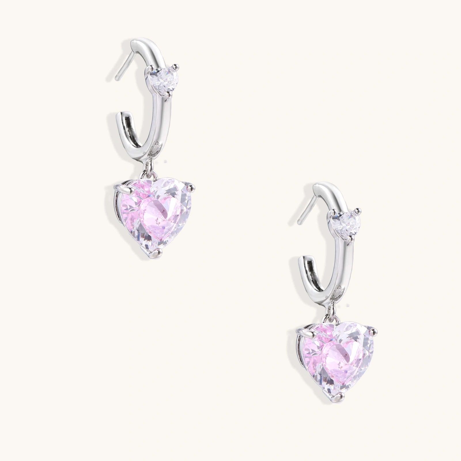 Silver huggie hoops with pink heart-cut zirconia pendants on white background, eco-friendly white gold plated brass