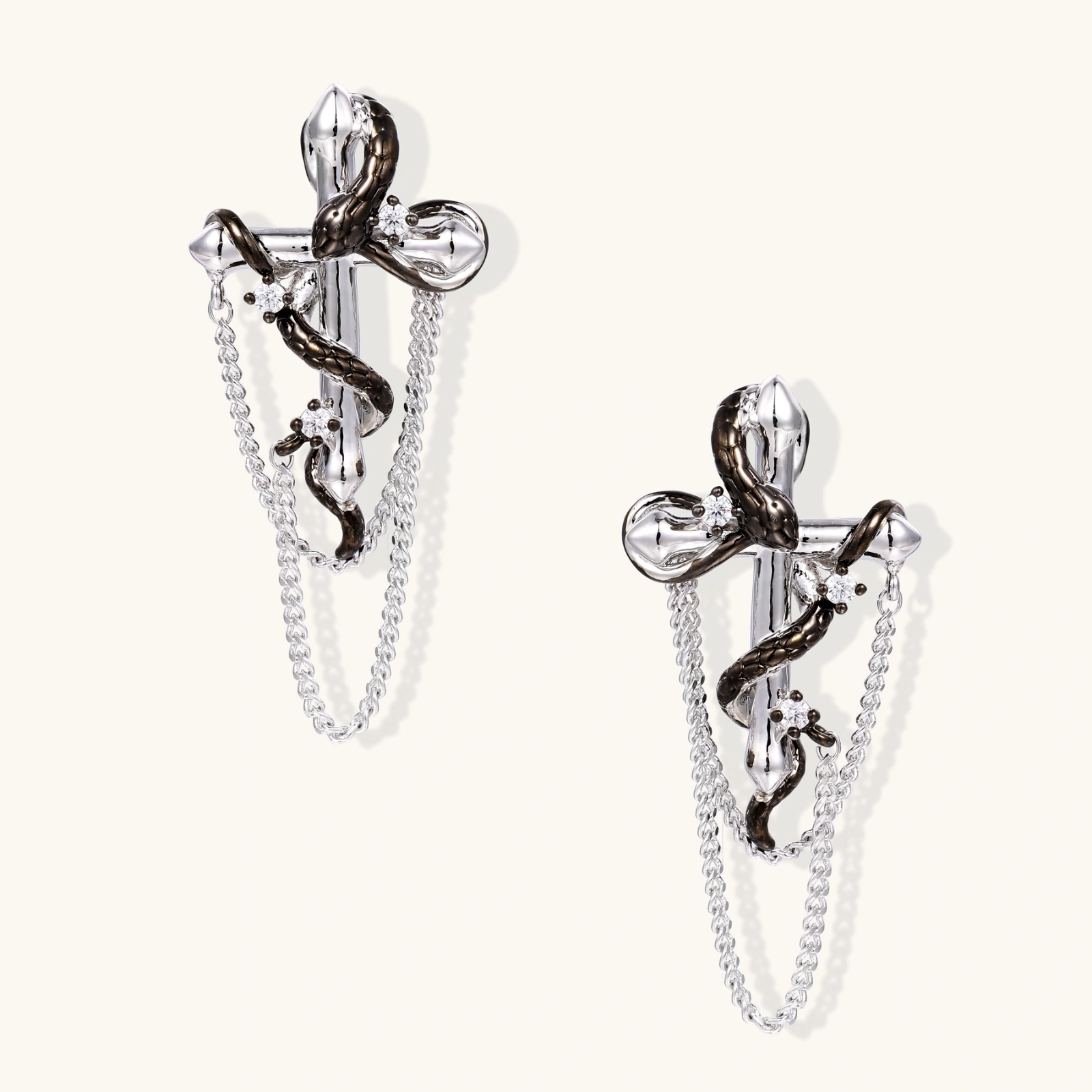 Silver dangle earrings with sculptural serpent coiled around cross shape and delicate chain accents on cream background