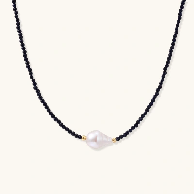 Delicate necklace with black blue sandstone beads and baroque pearl center on white background with gold accents.