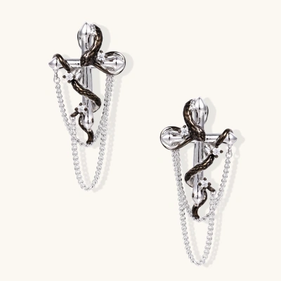Silver dangle earrings with sculptural serpent coiled around cross shape and delicate chain accents on cream background