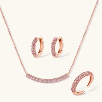 Rose gold jewelry set with pavé cubic zirconia stones including curved necklace, hoop earrings, and adjustable ring on cream background.