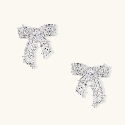 Elegant silver bow-shaped earrings with sparkling zirconia crystals and dangling ribbon details on cream background.