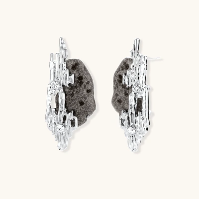 Sculptural silver earrings with dark volcanic stones, flowing metal design, and crystal accents on white background.