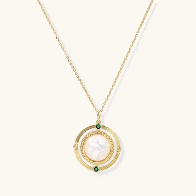 Gold plated necklace with a round white MOP pendant, textured gold ring, and two green zirconia stones on a cream background.