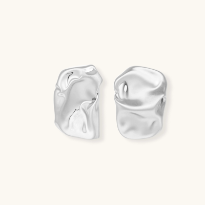 Pair of sculptural silver ear cuffs with organic fluid shapes and polished finish against cream background.