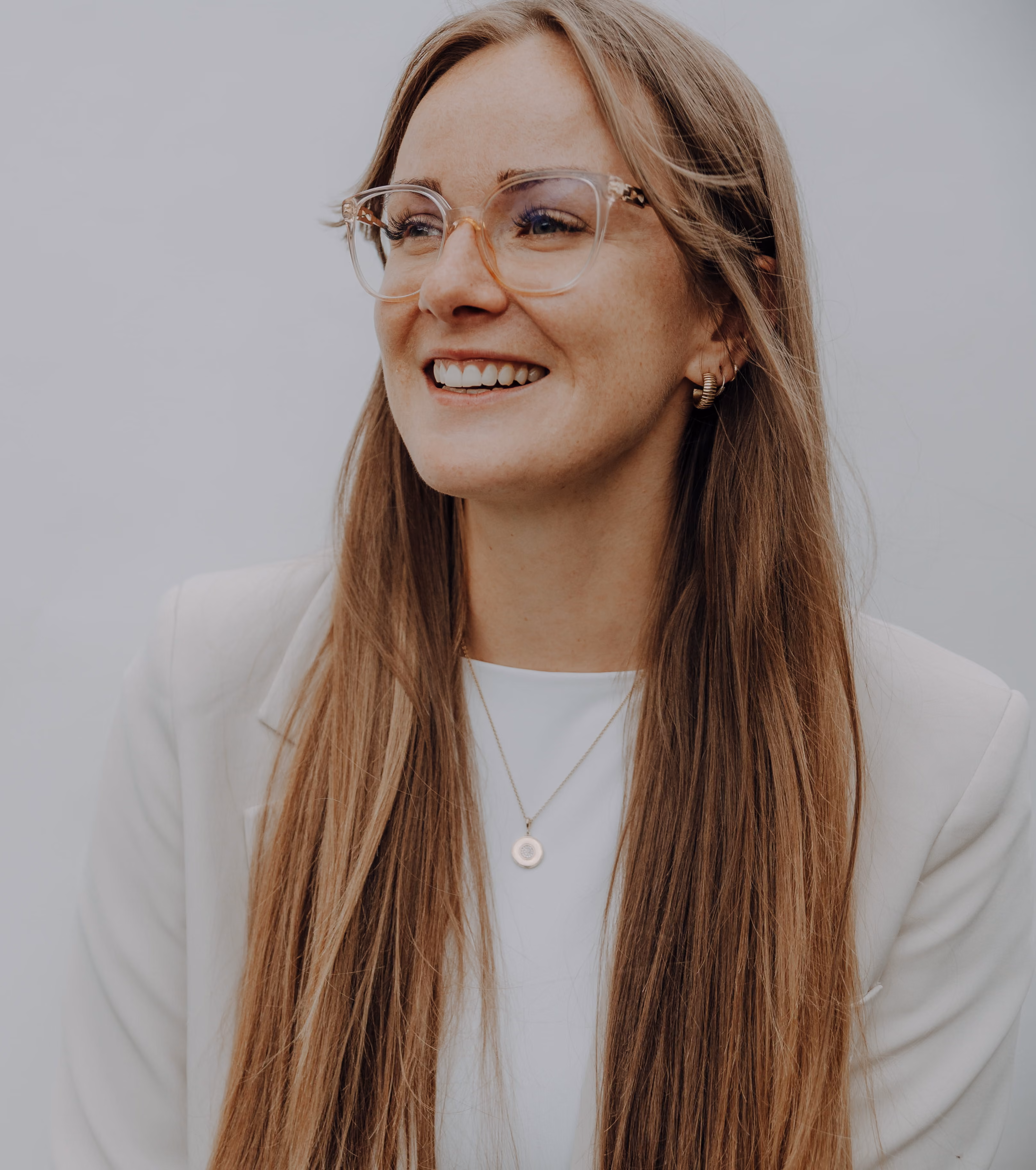 Porträt von Lorelle Dehnhard mit langen blonden Haaren, die klare Brille, weißen Blazer, weißen Oberteil und goldene Halskette trägt.