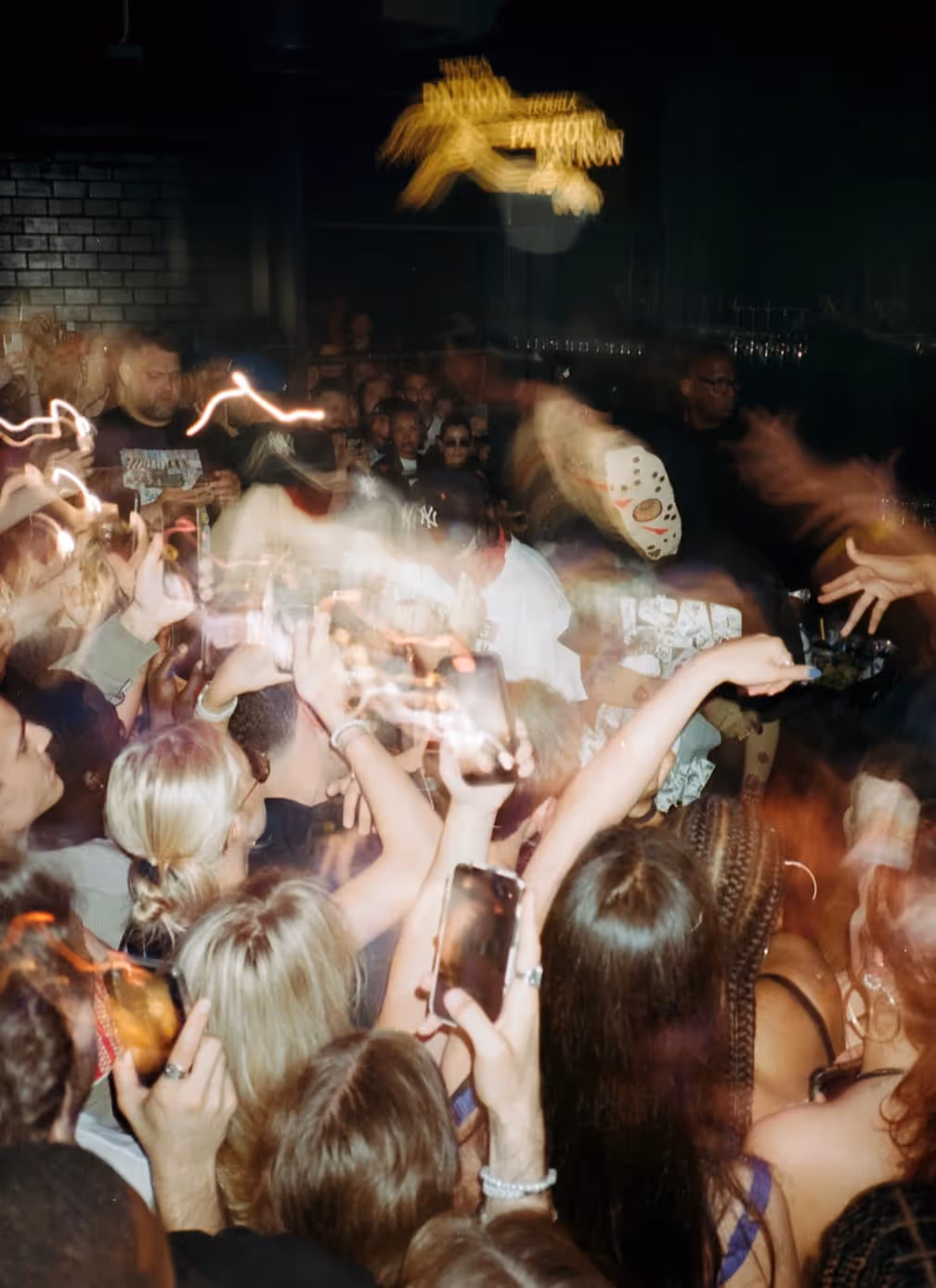 Crowd of people at a lively indoor event, many holding up phones capturing the moment, with motion blur and dim lighting.
