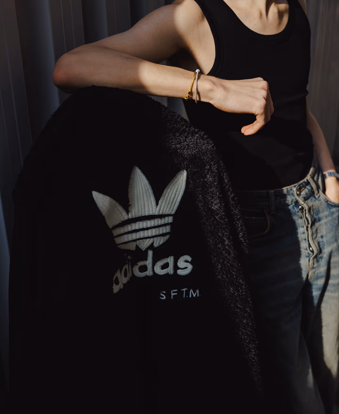 Person wearing a black tank top and jeans, holding a black Adidas jacket with white logo lettering.