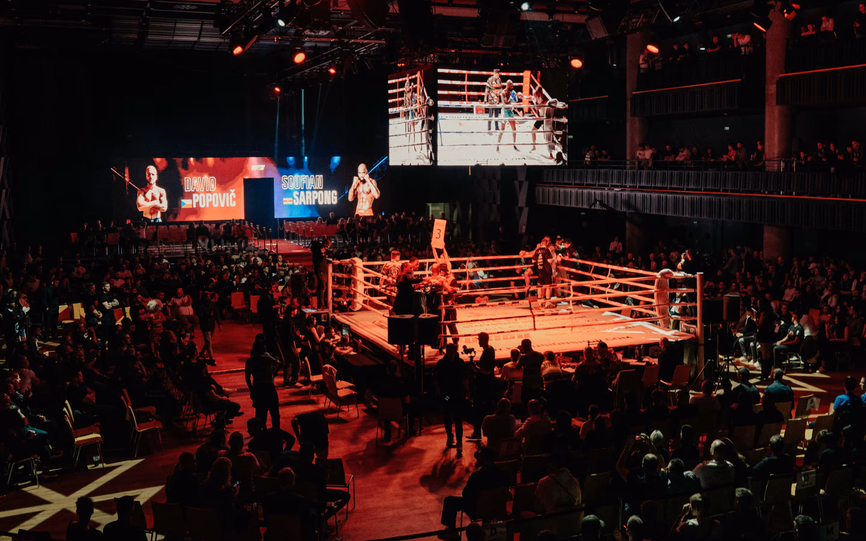 Boxing ring illuminated in red light with fighters and officials surrounded by a seated audience in a dark arena.