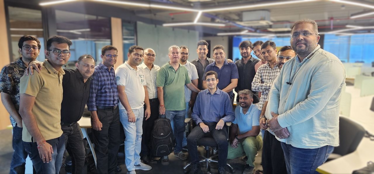 Group of 16 men posing together in an office setting, with one man seated in the center on an office chair.