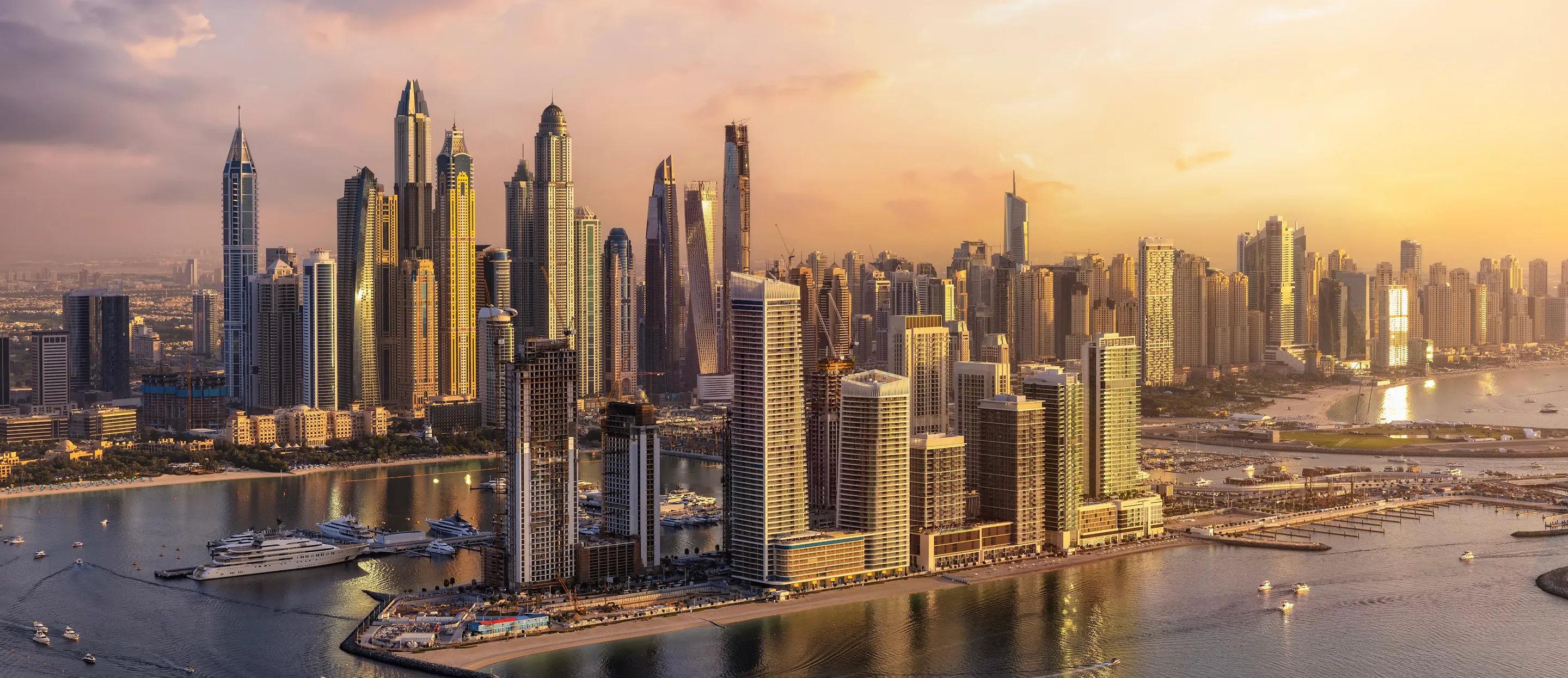 A sprawling city skyline with numerous modern skyscrapers reflecting golden sunset light along a waterfront with boats.